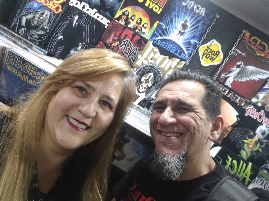 A smiling couple poses in front of a wall displaying band t-shirts and album art.
