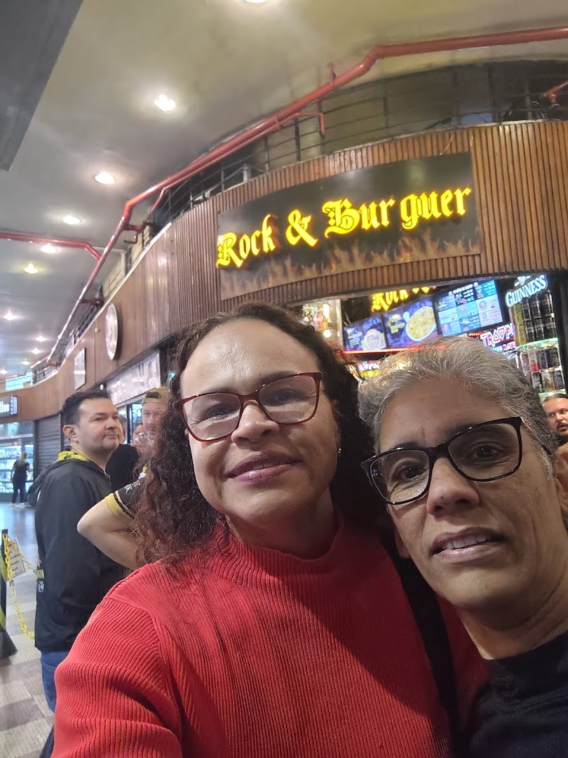 Two women pose for a selfie in front of a "Rock & Burguer" shop with other patrons in the background.