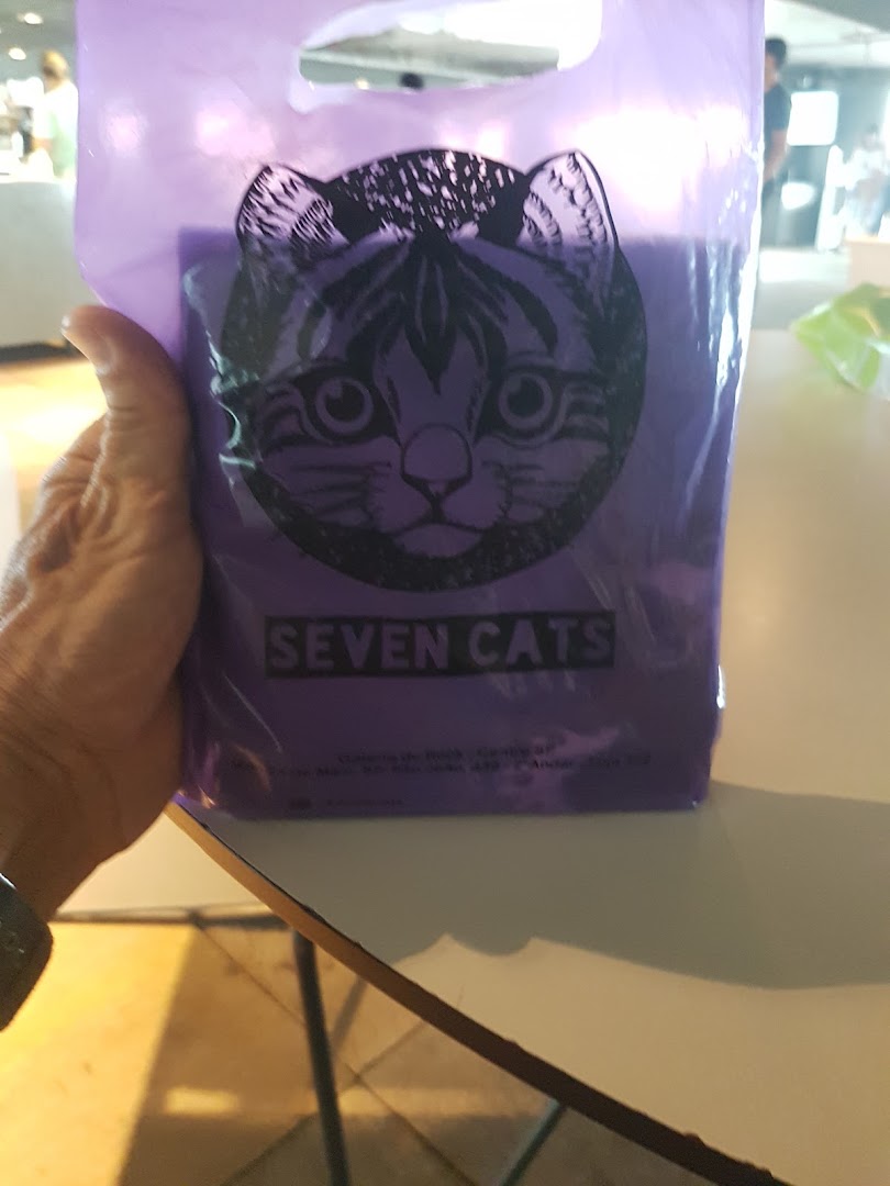 A hand holds a purple shopping bag with a cat logo and the text 'SEVEN CATS'.