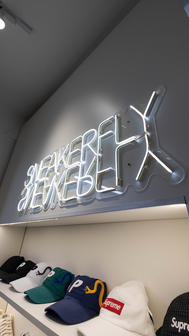 A neon sign for SNEAKERFLY above a display of various baseball caps.