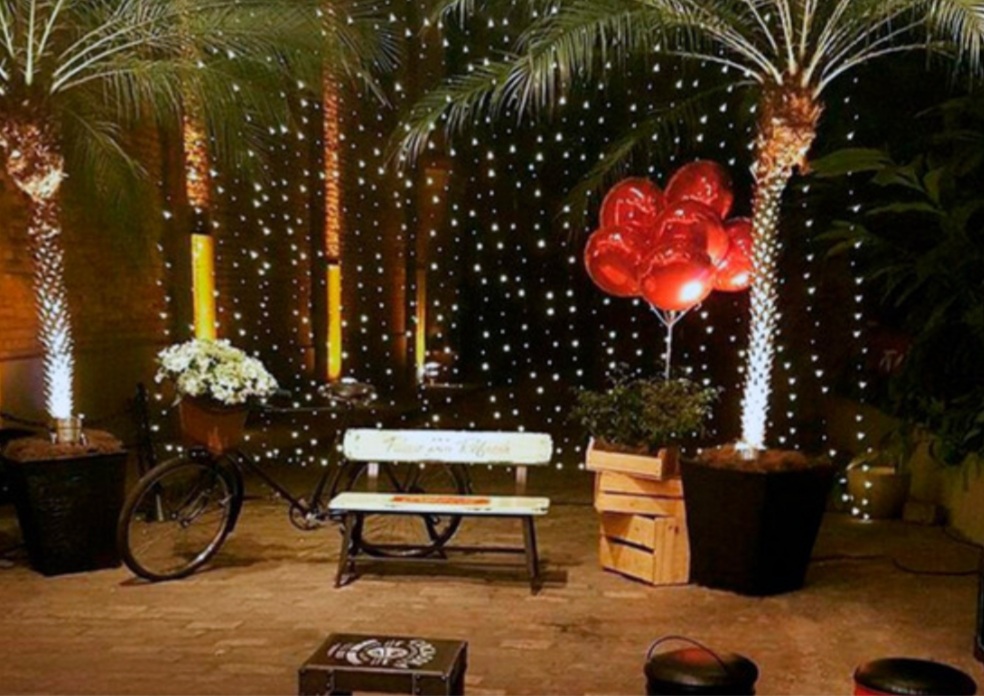 A festive outdoor scene with palm trees, fairy lights, red balloons, and a vintage bicycle.