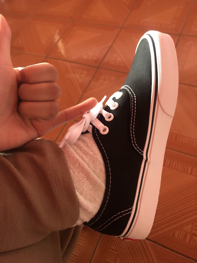 A person gives a thumbs up while wearing a black Vans sneaker with white laces.