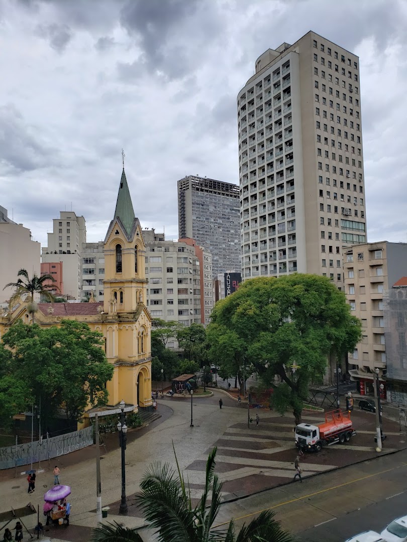 A city square with a church, tall buildings, trees, and people on a cloudy day.