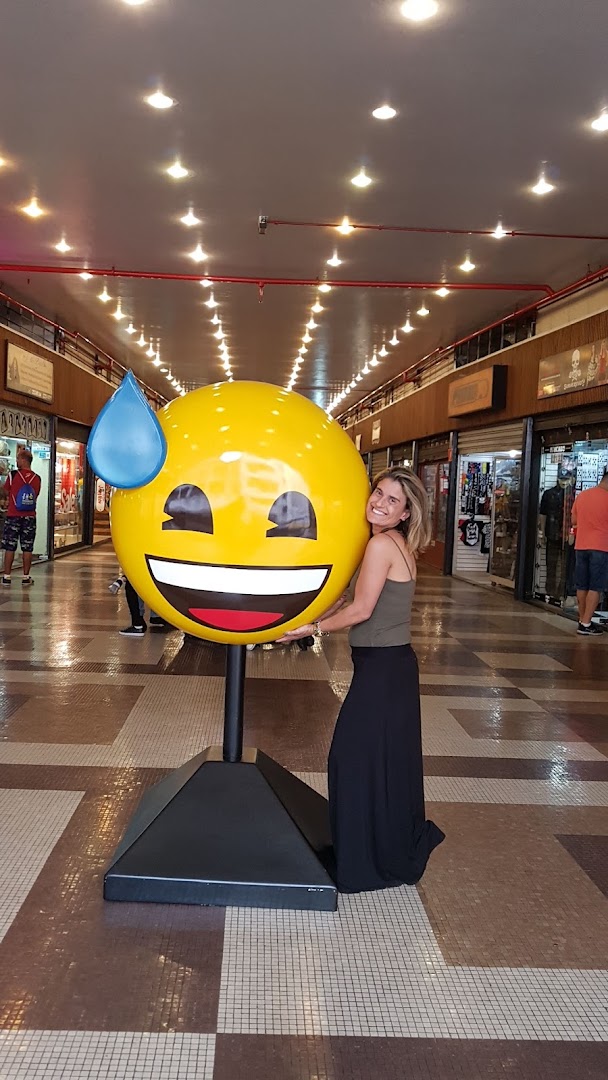 A woman poses with a large, sweating emoji statue in a brightly lit shopping arcade.