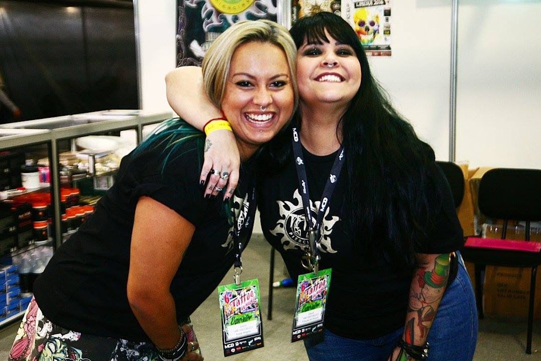 Two smiling women with piercings and tattoos, wearing black t-shirts with lanyards.