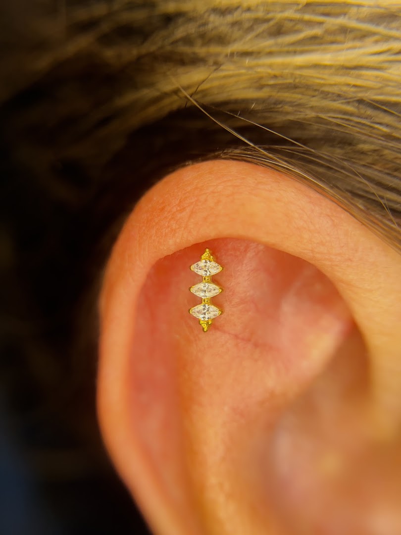 A close-up of a helix piercing with a gold barbell featuring three marquise-cut clear stones.