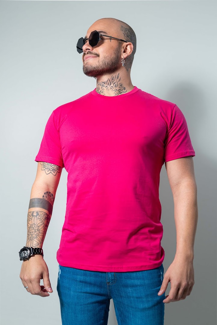 A man with tattoos and sunglasses wears a bright pink t-shirt and blue jeans.
