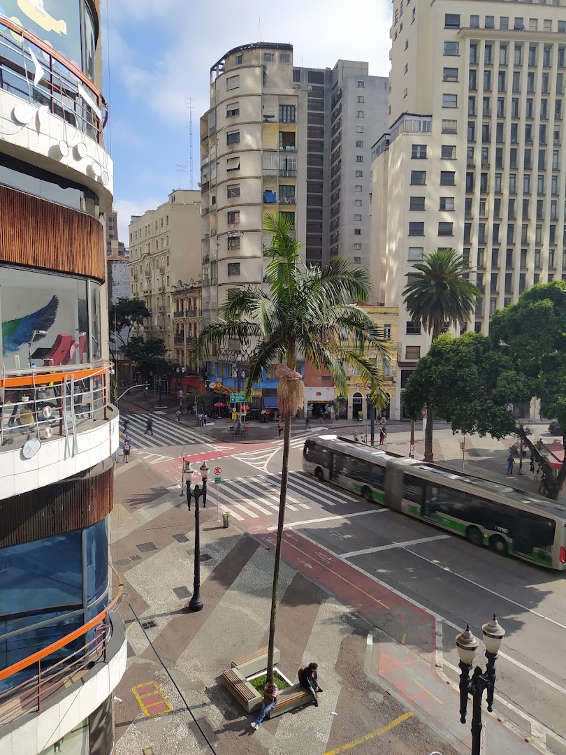 A street scene with buildings, a bus, palm trees, and people sitting on a bench.