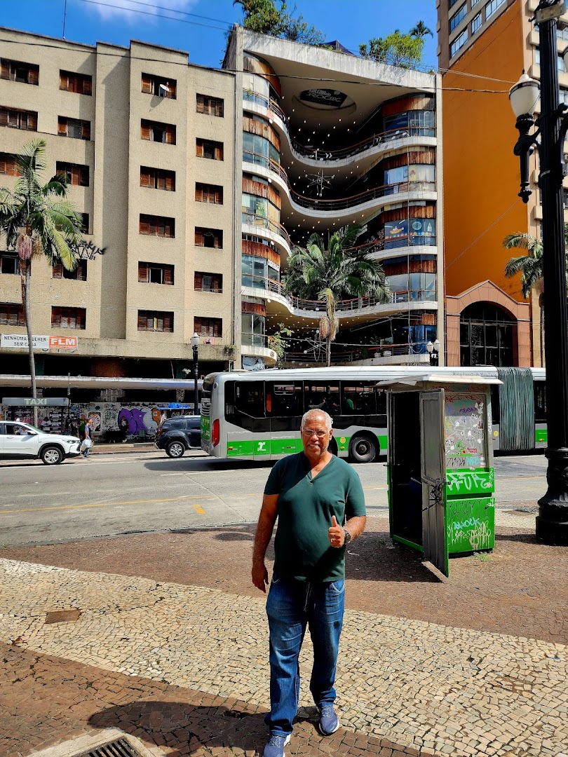 A man stands on a sidewalk in front of a distinctive curved building with a bus and cars passing by.