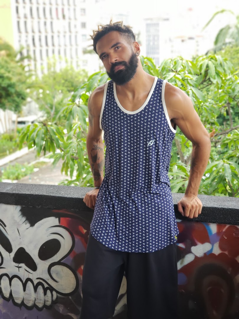 A man with dreadlocks and a beard poses wearing a patterned tank top and black shorts.