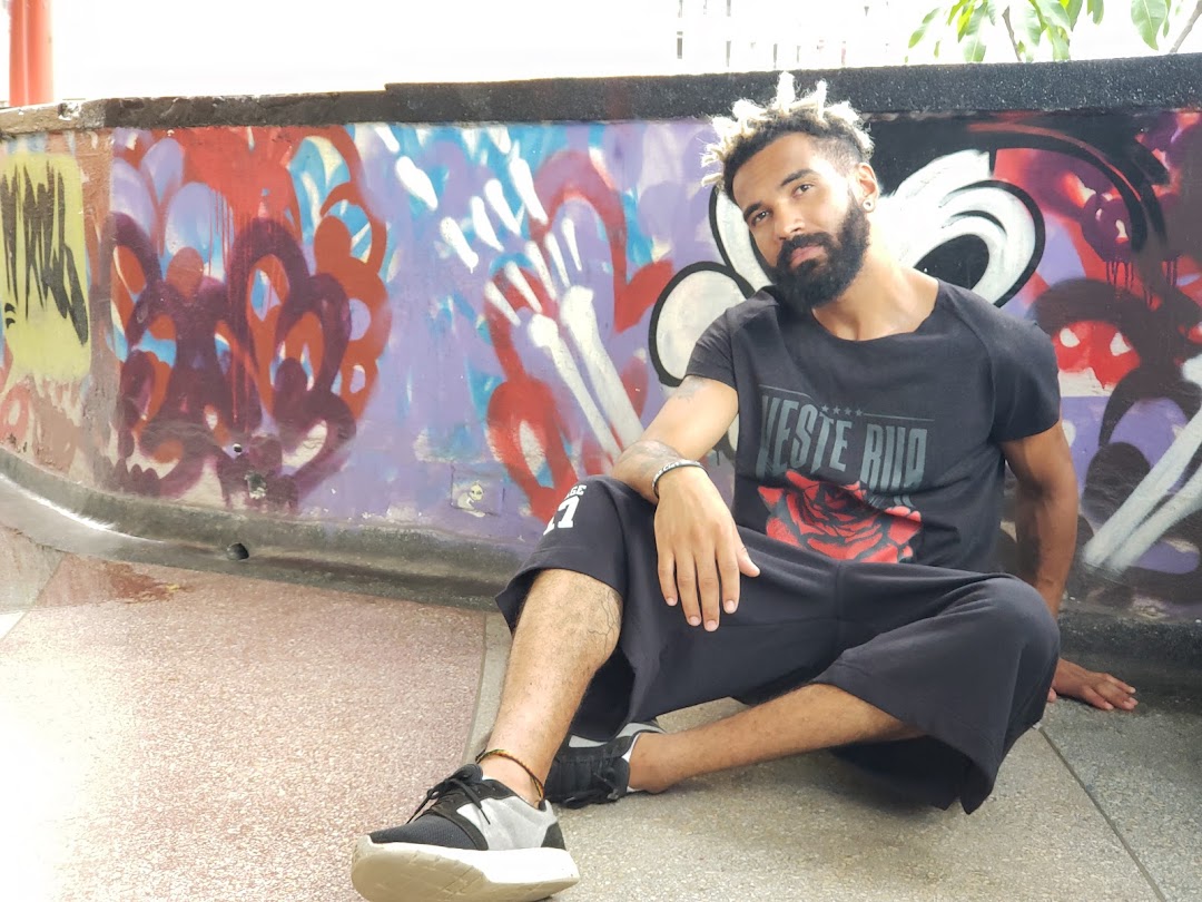 A man with dreadlocks and a beard sits in front of a graffiti-covered wall, wearing a black t-shirt.