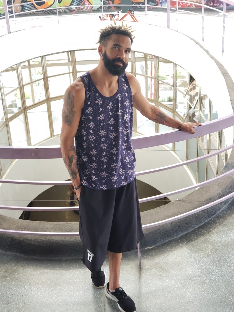 A man with dreadlocks and a beard poses in a patterned tank top and shorts.