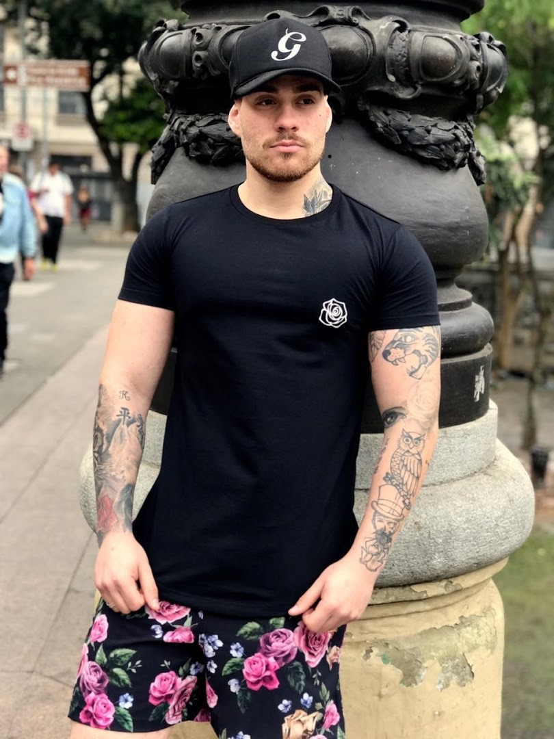 A man wearing a black t-shirt and floral shorts poses next to a stone pillar.