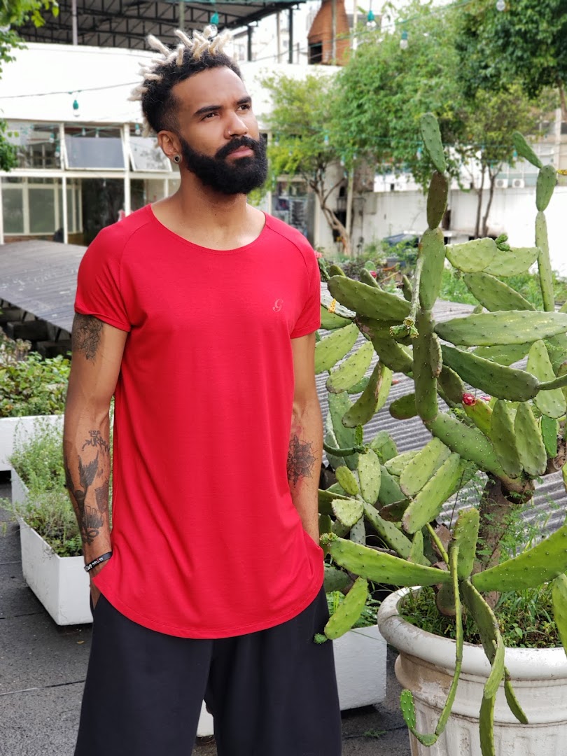 A man with dreadlocks and a beard wears a red t-shirt and black pants, posing outdoors near cacti.