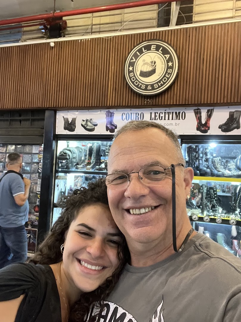 A smiling couple poses in front of a Vilela Boots & Shoes store displaying various footwear.