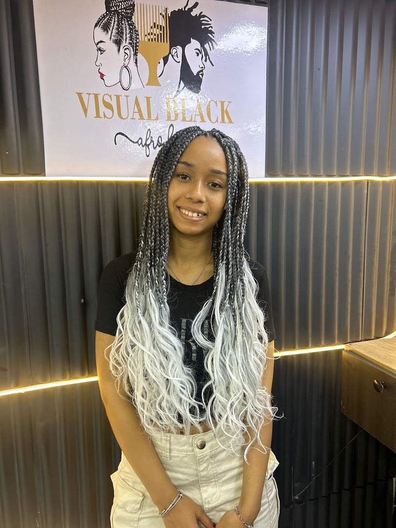 A smiling young woman with long, grey and white braided hair stands in front of the Visual Black salon logo.