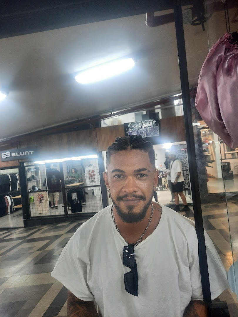 A man with braided hair and tattoos poses in front of a retail shop.