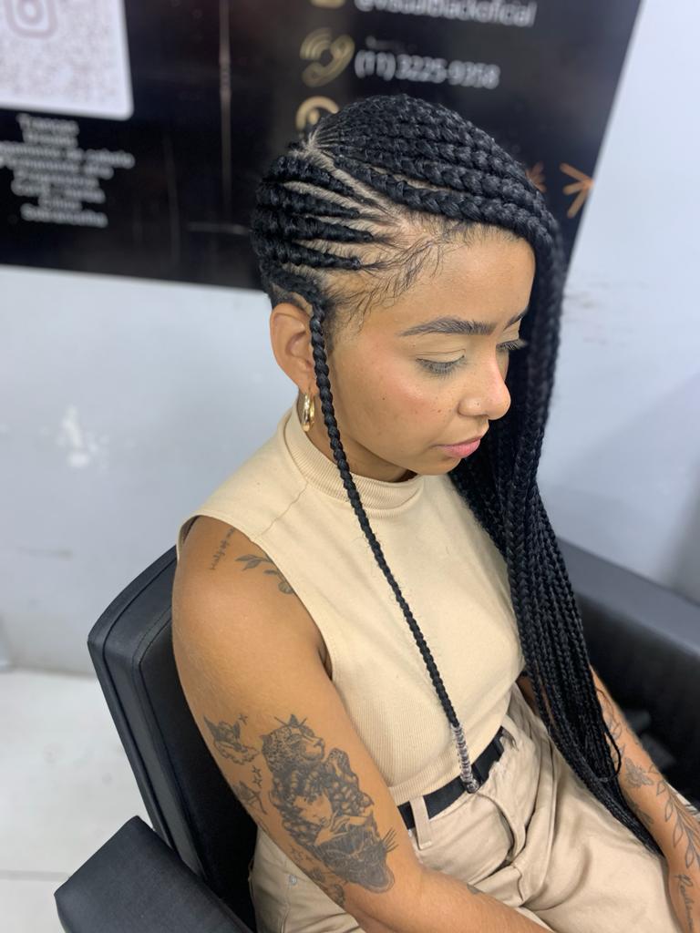A woman with intricate braided hair and tattoos sits in a salon chair.