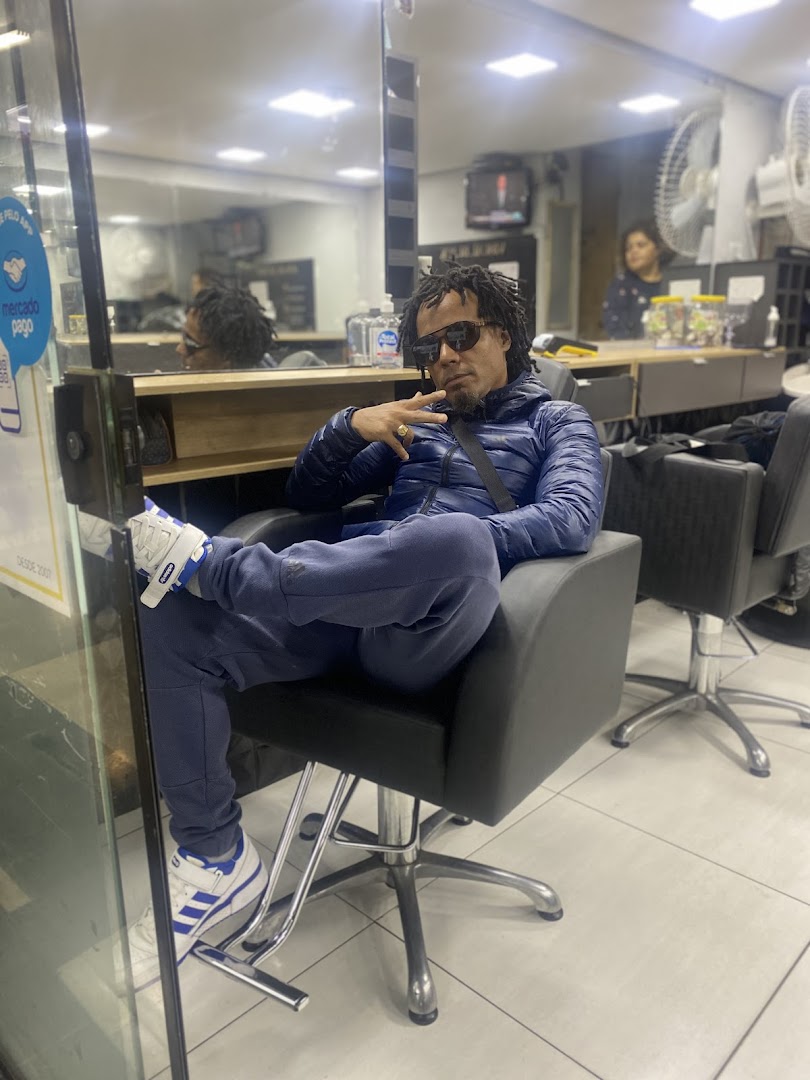 A man with dreadlocks sits in a barber chair, posing for the camera.