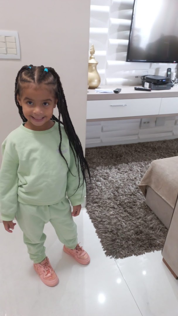 A young girl with braided hair smiles while wearing a light green tracksuit and pink sneakers.
