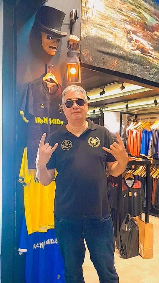 A man poses in a shop selling Iron Maiden merchandise, with band shirts displayed.