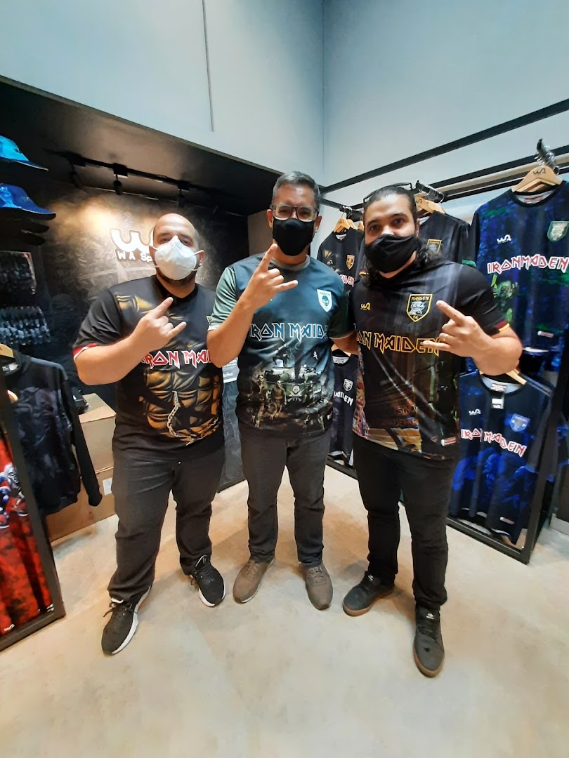Three men wearing Iron Maiden t-shirts pose in a store with merchandise displayed.