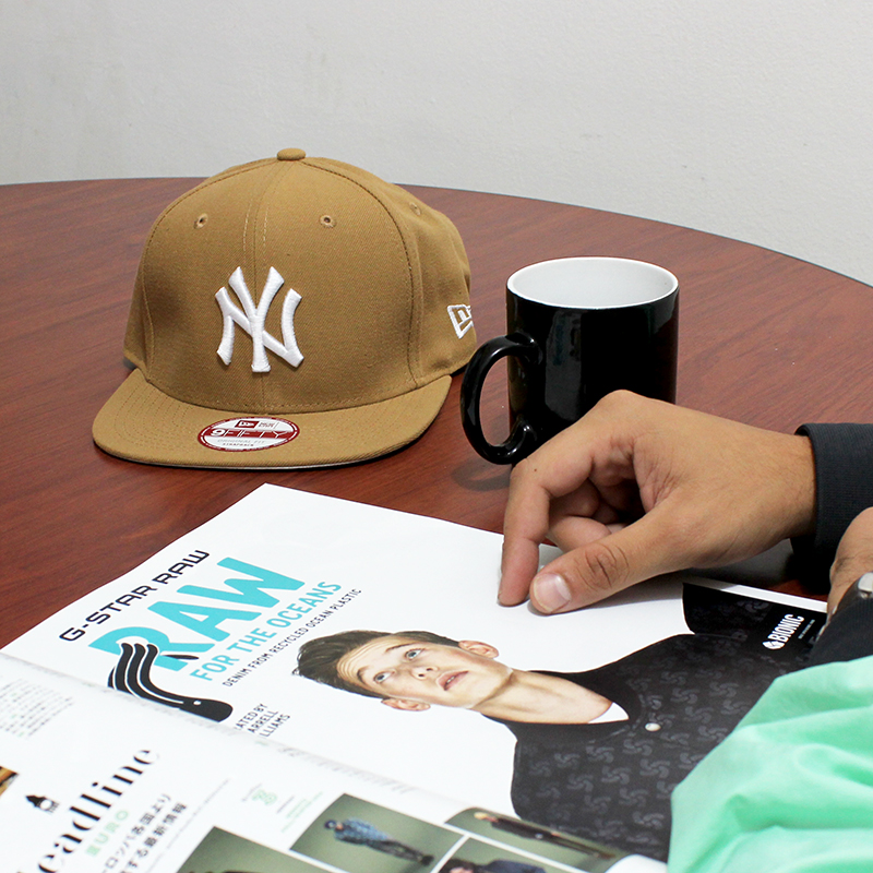 A New Era Yankees cap and a magazine are on a table with a mug and a hand.