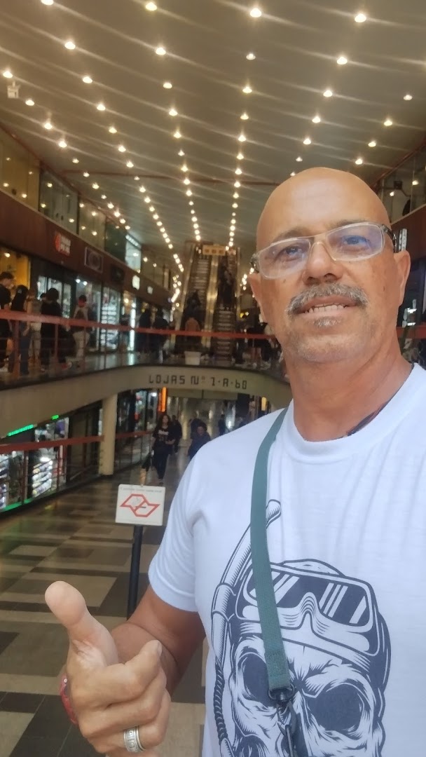 A man gives a thumbs up in a shopping mall with escalators and shops visible.