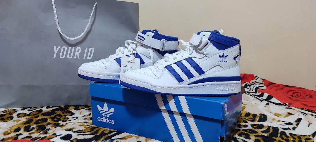 White and blue Adidas Forum Mid sneakers displayed with their box and a shopping bag.